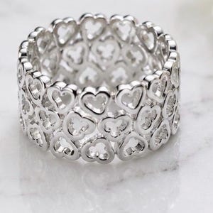 Puede incluir: Un anillo de banda ancha de plata con un patrón calado repetitivo de formas de corazón interconectadas. El metal tiene un acabado pulido y reflectante, y el diseño consta de tres filas horizontales de corazones que forman una estructura.