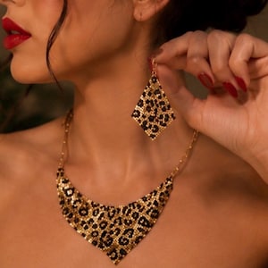 Puede incluir: Un collar de tono dorado y un pendiente a juego en forma de diamante con un estampado de leopardo en negro y oro. El conjunto de joyería está elaborado con una textura fina similar a una malla, mostrando un diseño de manchas animales.