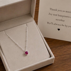 Puede incluir: Un collar de cadena plateada con un pequeño colgante de piedra roja en forma de corazón, presentado en un joyero beige. Junto a la caja hay una tarjeta con el texto: Thank you so much, Joy and happiness of ayerday, We'll always be by you, con un icono de corazón.