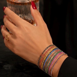 Puede incluir: Un conjunto de siete pulseras de tenis finas y flexibles con pequeñas piedras redondas y brillantes en un espectro vibrante de colores que incluye negro, rojo, amarillo, azul, verde, rosa y morado, usadas juntas como un accesorio apilado.