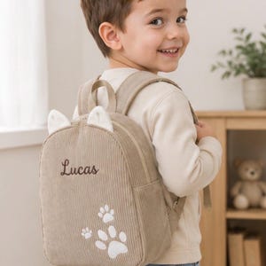 Puede incluir: Una mochila de pana beige con dos pequeñas orejas blancas en la parte superior, el nombre Lucas bordado en letra cursiva marrón oscuro y un gráfico de huella blanca en la parte inferior frontal, con correas ajustables y cierre de cremallera.