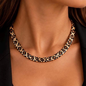 Puede incluir: Un collar tipo gargantilla en tono dorado con un patrón repetitivo de manchas de leopardo negras y marrones. El collar tiene un diseño segmentado, plano y liso que se ajusta al cuello, creando una banda continua de estampado animal.