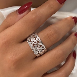 Puede incluir: Un anillo de banda ancha en plata con un intrincado patrón calado de pequeños corazones interconectados que se repiten. El metal tiene un acabado pulido y reflectante, creando un diseño continuo y texturizado alrededor del dedo.