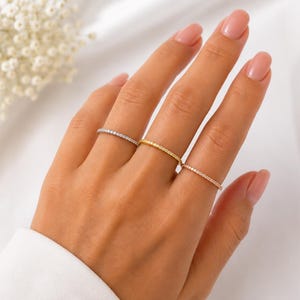 Puede incluir: Tres anillos finos y delicados en los dedos. Un anillo es de tono plateado, otro de tono dorado y otro de tono oro rosa. Cada anillo presenta una hilera continua de pequeñas piedras claras y brillantes a lo largo de toda la circunferencia.