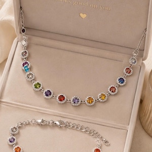Puede incluir: Un conjunto de collar y pulsera plateados con piedras preciosas redondas y multicolores rodeadas de pequeñas piedras transparentes. Las joyas se presentan en una caja beige con el texto "Good energy looks good on you" y un pequeño corazón dorado.