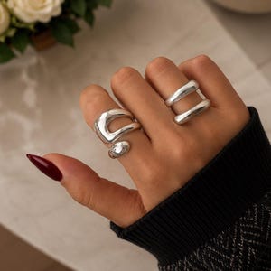 Puede incluir: Dos anillos de plata con superficies lisas y pulidas. Un anillo presenta una forma orgánica abierta e irregular en el dedo índice, mientras que el otro es un anillo de doble banda con dos barras de plata paralelas en el dedo medio.