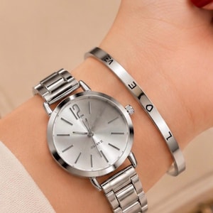 May include: A silver-toned analogue wristwatch with a metallic link band and a round face featuring the number 12 and stick markers, paired with a thin, rigid silver-toned bangle bracelet engraved with the word LOVE in black capital letters.