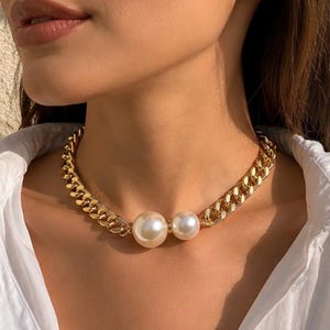 Puede incluir: Un collar de cadena de eslabones gruesos en tono dorado con dos perlas blancas grandes, redondas y lisas situadas en el centro. El collar descansa sobre el cuello, mostrando un acabado metálico pulido y un diseño de eslabones clásico con cierre.