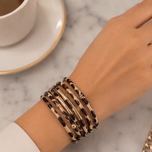 Puede incluir: Un conjunto de cinco pulseras finas apiladas con un estampado de leopardo en marrón y negro. Las pulseras están adornadas con pequeñas cuentas metálicas doradas, lisas, de forma cilíndrica y esférica, intercaladas en los aros.