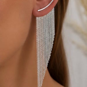 Puede incluir: Un pendiente tipo ear crawler de plata con una barra metálica curva que se asienta sobre el lóbulo, con múltiples cadenas de plata finas y largas que cuelgan en cascada con longitudes graduadas, creando un efecto de flecos.