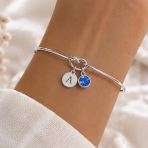 Puede incluir: Una pulsera rígida de tono plateado con un diseño de nudo central. Dos pequeños dijes circulares cuelgan del nudo: un disco plateado grabado con la letra mayúscula A y un dije circular con una piedra preciosa de color azul brillante.