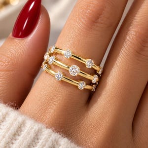 Puede incluir: Un anillo de oro de tres bandas que presenta múltiples piedras pequeñas, redondas, transparentes y facetadas, engastadas a intervalos a lo largo de cada banda fina. El diseño consta de tres bandas doradas paralelas y separadas con piedras.