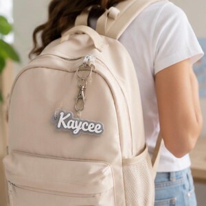 Puede incluir: Una mochila beige con un llavero personalizado de acrílico gris y blanco que muestra el nombre Kaycee en letra cursiva. El llavero está sujeto al tirador de la cremallera superior mediante un mosquetón de metal de tono plateado.