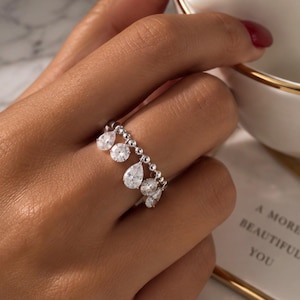 Puede incluir: Un anillo de tono plateado con una banda de cuentas y piedras transparentes alternadas en forma de pera y redondas que cuelgan de la base. El diseño crea un efecto colgante a lo largo del dedo, mostrando múltiples cristales blancos facetados.