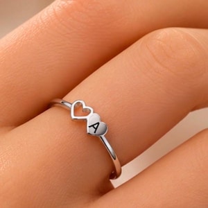 Puede incluir: Un anillo de metal en tono plateado con dos pequeños elementos en forma de corazón en la banda. Un corazón es un contorno abierto, mientras que el otro es un corazón sólido grabado con la letra mayúscula A en una fuente negra y clara.