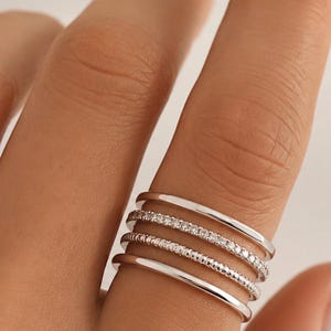 Puede incluir: Un conjunto de cuatro anillos de tono plateado usados juntos en un dedo. El grupo incluye dos bandas lisas y simples, una banda con una hilera de pequeñas piedras transparentes y una banda con una superficie texturizada y granulada, con acabado pulido.