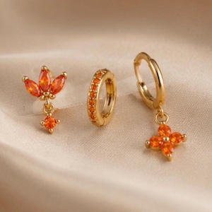 Puede incluir: Un conjunto de tres pendientes dorados con piedras de color naranja brillante. Un pendiente tiene un racimo de corte marquesa con una piedra redonda colgante, otro es un aro pequeño con piedras naranjas incrustadas y otro es un aro con flor.
