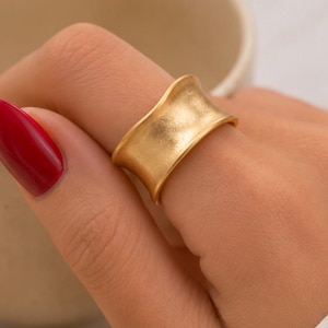 Può includere: Un ampio anello dorato con finitura opaca spazzolata e un design a fascia concava e curva. L'anello presenta una superficie liscia e leggermente testurizzata che cattura la luce, creando sottili riflessi lungo i bordi della fascia.