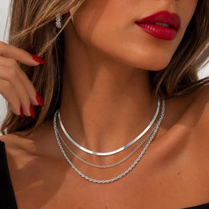 Puede incluir: Un conjunto de collares plateados en capas que consta de tres cadenas distintas. La capa superior es una cadena de espiga plana y lisa, seguida de una delicada cadena de eslabones en el medio y una cadena de cuerda texturizada en la parte inferior.