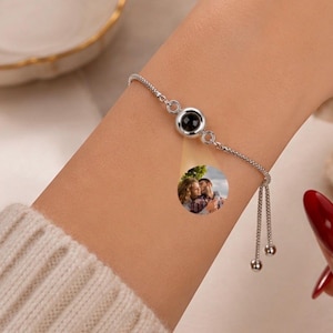 Puede incluir: Una pulsera de cadena en tono plateado con un dije circular central que proyecta una pequeña fotografía circular en color de dos personas abrazándose. La pulsera tiene una cadena de eslabones delicada y dos extremos colgantes con pequeñas cuentas esféricas.