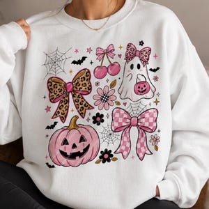 Puede incluir: Una sudadera blanca de cuello redondo con un estampado gráfico de una calabaza rosa, un fantasma con un cubo de calabaza, cerezas y varios lazos con estampado de leopardo y cuadros rosas, decorada con telarañas, flores y pequeños murciélagos.