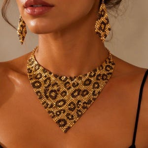 Puede incluir: Un collar de malla metálica dorada y pendientes colgantes a juego en forma de diamante, ambos con un estampado de leopardo en marrón oscuro y dorado. El collar tiene un diseño ancho en forma de V que descansa sobre la clavícula.
