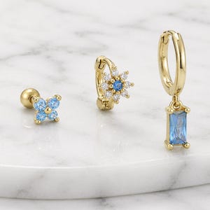 Puede incluir: Un conjunto de tres pendientes dorados con piedras azul claro y transparentes. Uno es un pendiente de botón de cuatro pétalos, otro es un aro con un dije de flor con piedra central azul, y el tercero es un aro con un colgante rectangular azul.
