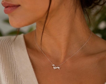 Op de afbeelding: Een zilverkleurige ketting met een kleine, platte hanger in de vorm van een teckel. De hanger is bevestigd aan een fijne, dunne ketting die om de nek rust. Het ontwerp is eenvoudig, metaalachtig en gepolijst met een gladde afwerking.