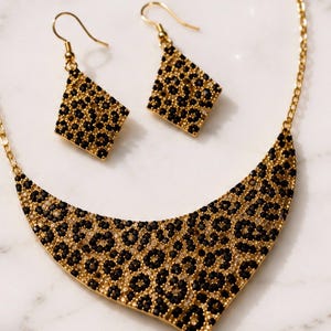 Puede incluir: Un collar dorado y un par de pendientes a juego, ambos con un estampado de leopardo compuesto por pequeños cristales negros y transparentes sobre una base de metal dorado. El collar tiene una forma ancha y curva de media luna.