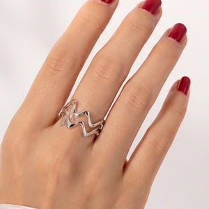 Puede incluir: Un anillo de tono plateado que presenta un patrón de zigzag u onda de doble capa. La banda está construida con dos líneas metálicas paralelas e interconectadas que crean un diseño continuo, rítmico y angular alrededor de la circunferencia.