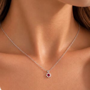 Puede incluir: Un collar de cadena en tono plateado con un colgante central que presenta una piedra preciosa redonda de color rojo intenso, rodeada por un halo de pequeñas piedras claras y brillantes dispuestas en forma de flor.