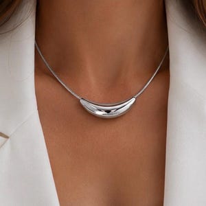 Puede incluir: Un collar de tono plateado que presenta un colgante liso y curvo en forma de media luna, suspendido de una cadena de serpiente fina y flexible. La superficie de metal pulido refleja la luz, creando un aspecto brillante y metálico.
