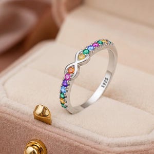 May include: A silver-toned ring featuring an infinity symbol centerpiece and a band set with small, round, multi-colored gemstones in shades of green, yellow, orange, pink, purple, and blue. The interior of the band is stamped with the text S925.