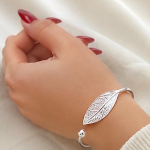May include: A silver-toned cuff bracelet featuring a detailed leaf-shaped centerpiece with an intricate, textured vein pattern. The open-ended design terminates in small, smooth spherical beads at each end, creating a polished metallic look.