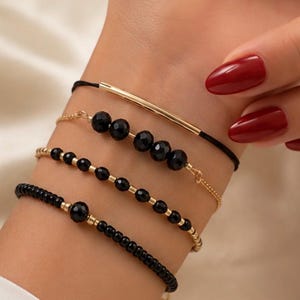 Puede incluir: Un conjunto de cuatro pulseras usadas juntas. Una presenta un cordón negro fino con una barra de metal en tono dorado. Otra es una cadena dorada con cuatro cuentas negras grandes facetadas. La tercera es una cadena dorada con cuentas negras pequeñas. La cuarta es una pulsera de cuentas negras con una cuenta negra más grande.