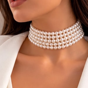 Puede incluir: Un collar tipo gargantilla de varias hileras que presenta cuatro filas de perlas uniformes, redondas y de color crema. El collar se ajusta al cuello, mostrando un acabado consistente, suave y brillante en cada perla individual.
