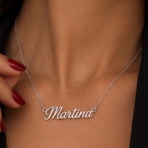 Puede incluir: Un collar de tono plateado con un colgante de placa con el nombre Martina escrito en cursiva. El colgante está unido a una cadena fina y delicada de tono plateado que descansa alrededor del cuello, mostrando un acabado metálico pulido.
