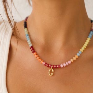 Puede incluir: Un collar que presenta una hilera de pequeñas cuentas multicolores en tonos rojo, naranja, amarillo, azul y morado. Un colgante de metal dorado con forma de letra mayúscula C cuelga del centro de la cadena de cuentas del collar.