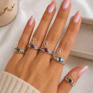 Puede incluir: Cinco anillos de tono plateado con grabados en forma de estrella y varias piedras preciosas de colores. El anillo uno tiene una estrella verde, el dos una piedra redonda morada, el tres un corazón rosa, el cuatro una piedra azul rectangular.