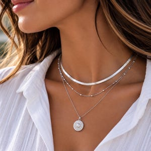 May include: A layered silver necklace set consisting of three distinct chains. The top layer is a flat herringbone chain, the middle is a delicate beaded chain, and the bottom is a thin cable chain featuring a round, textured medallion pendant.