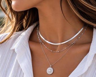 May include: A layered silver necklace set consisting of three distinct chains. The top layer is a flat herringbone chain, the middle is a delicate beaded chain, and the bottom is a thin cable chain featuring a round, textured medallion pendant.