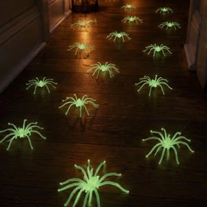 May include: A collection of small, glow-in-the-dark spider figurines scattered across a dark wooden floor. Each spider features eight curved legs and a luminous, pale green appearance that stands out against the dark surface of the floor.