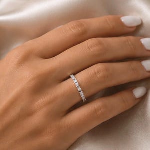 Puede incluir: Un anillo fino de tono plateado que presenta una hilera continua de pequeñas piedras redondas y transparentes en engaste de garras. El anillo tiene un acabado metálico pulido y un diseño uniforme que rodea toda la circunferencia de la banda.