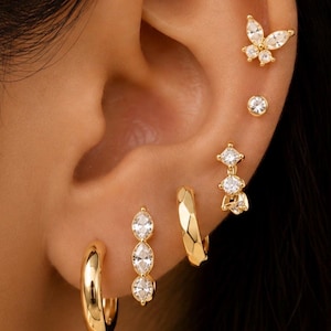 Puede incluir: Un conjunto de pendientes dorados en múltiples perforaciones, que incluye un pendiente en forma de mariposa con piedras claras, un pequeño pendiente redondo de cristal, un dije de cristal colgante, un aro facetado y un pendiente de tres piedras.