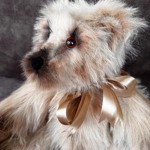 May include: A handmade teddy bear with a light brown and cream fur. The bear has a black nose, brown eyes, and a gold satin ribbon tied around its neck. The bear's fur is soft and fluffy, giving it a vintage look.