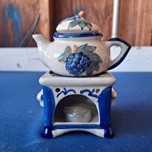 May include: A ceramic teapot warmer set featuring a cream-colored teapot with a blue grape and green leaf motif, resting on a matching square base with blue trim and a central opening containing a small white tea light candle for heating.
