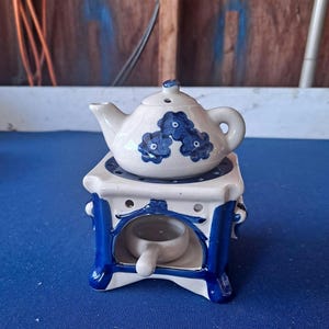 May include: A ceramic teapot with a cream-colored glaze and blue floral patterns sits atop a square, blue and cream-colored stove-shaped warmer. The warmer features a small, matching cream-colored ceramic bowl with a handle tucked inside its base.