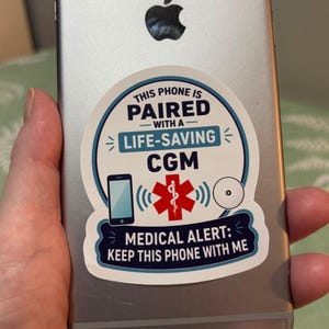 May include: A circular white sticker on a silver smartphone featuring blue text that reads THIS PHONE IS PAIRED WITH A LIFE-SAVING CGM and MEDICAL ALERT: KEEP THIS PHONE WITH ME, with a red medical symbol and icons of a phone and a sensor.