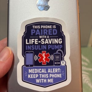 May include: A rectangular sticker on a silver smartphone with a dark blue background and white text reading THIS PHONE IS PAIRED WITH A LIFE-SAVING INSULIN PUMP and MEDICAL ALERT: KEEP THIS PHONE WITH ME, featuring a red medical symbol and icons.