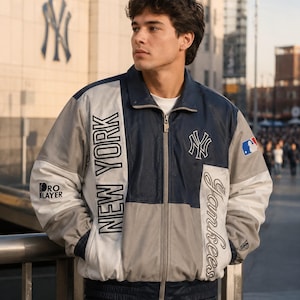 May include: A color-blocked zip-up jacket featuring navy blue, grey, and white panels. The front displays the text NEW YORK and Yankees, with a New York Yankees logo on the chest, a Pro Layer logo on the sleeve, and a Major League Baseball patch.